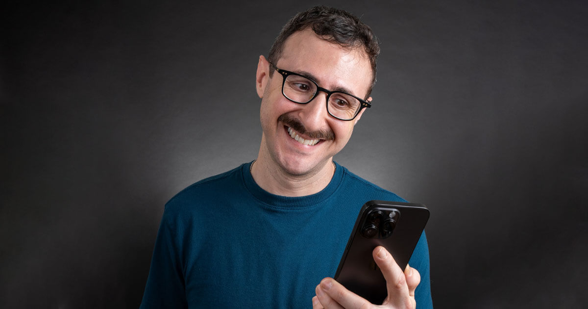 Dann looking at his black iPhone 14 Pro Max and smiling awkwardly. Studio photography, teal shirt with a black background.